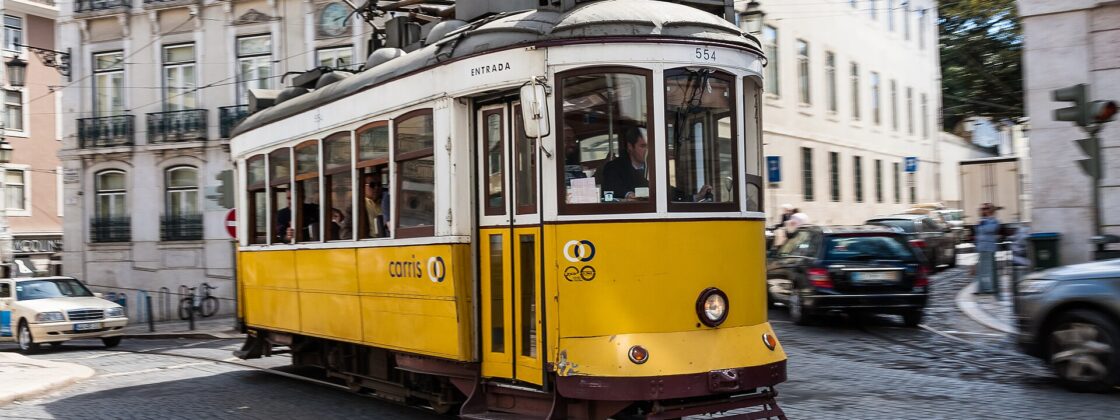 Yellow Carris Tram 28 turns along cobbled tracks in Lisbon, passing balconied buildings with passengers aboard.