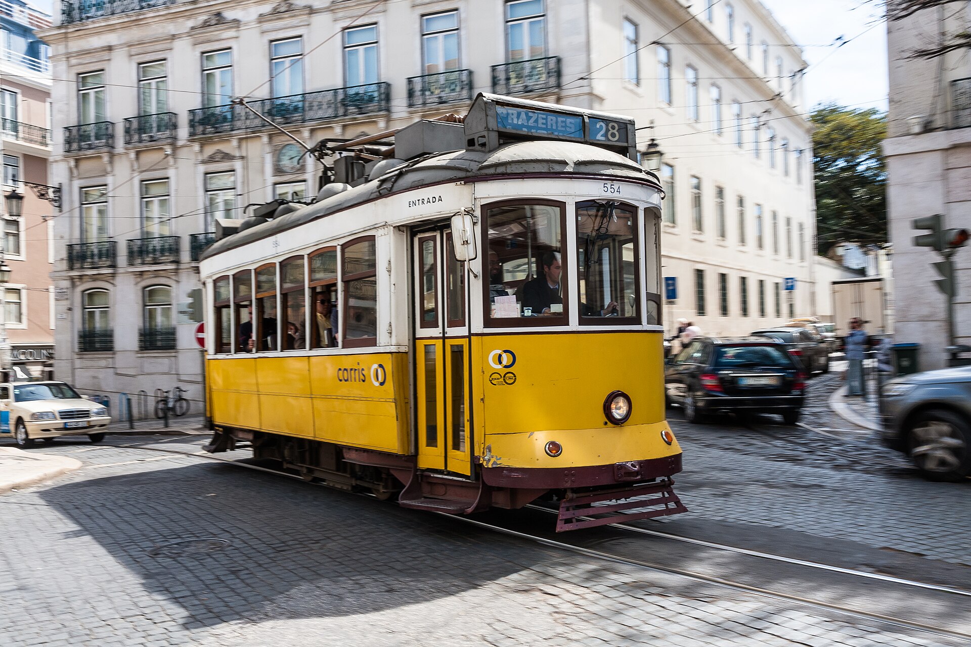 Yellow Carris Tram 28 turns along cobbled tracks in Lisbon, passing balconied buildings with passengers aboard.