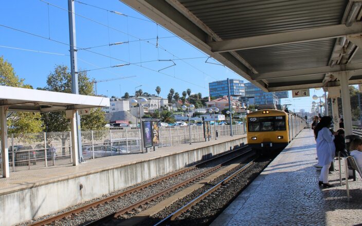 Yellow fronted CP suburban train at Cascais station, with passengers waiting beneath the platform canopy in bright sunshine.