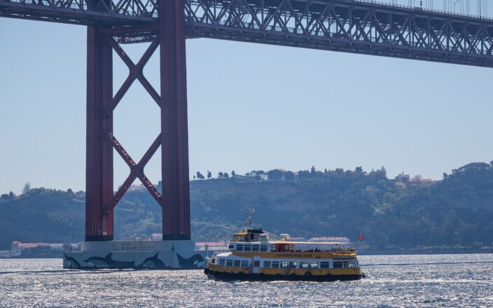 A yellow sightseeing boat carries passengers beneath Lisbon’s 25 de Abril Bridge on the sparkling Tagus River.