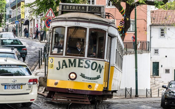 Yellow Tram 28 rounds a sloping Lisbon street beneath leafy trees, with passengers behind the Estrela destination board.