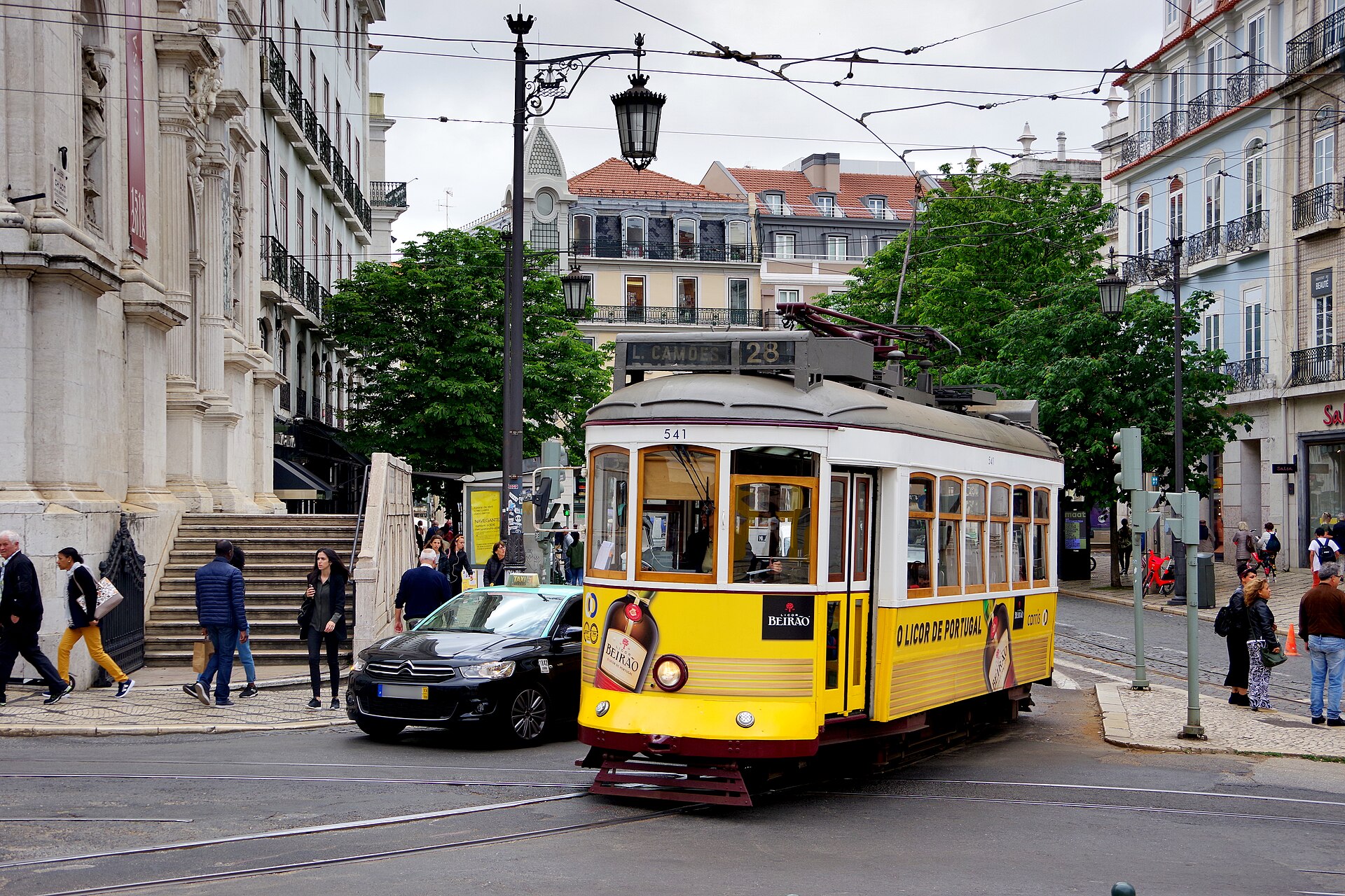 Yellow Tram 28 turns beside a Chiado church in Lisbon, with pedestrians, leafy trees and a taxi nearby.